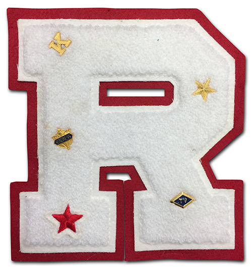 A white felt letter R is set against a red felt background. On the letter are five small pins, a letter K, a gold star, a red star, a badge with the word kindness printed on it, and a diamond with the letters TV in the center. 